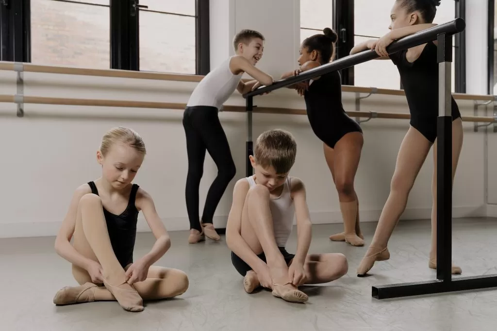 a few children in a dance studio, two of the children are sat on the floor while putting on their skin tone matching dance socks