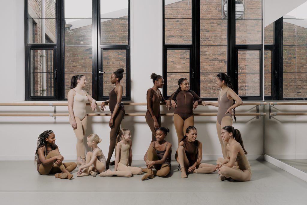 A diverse group of dancers stood around in a dance class next to the Ballet barre