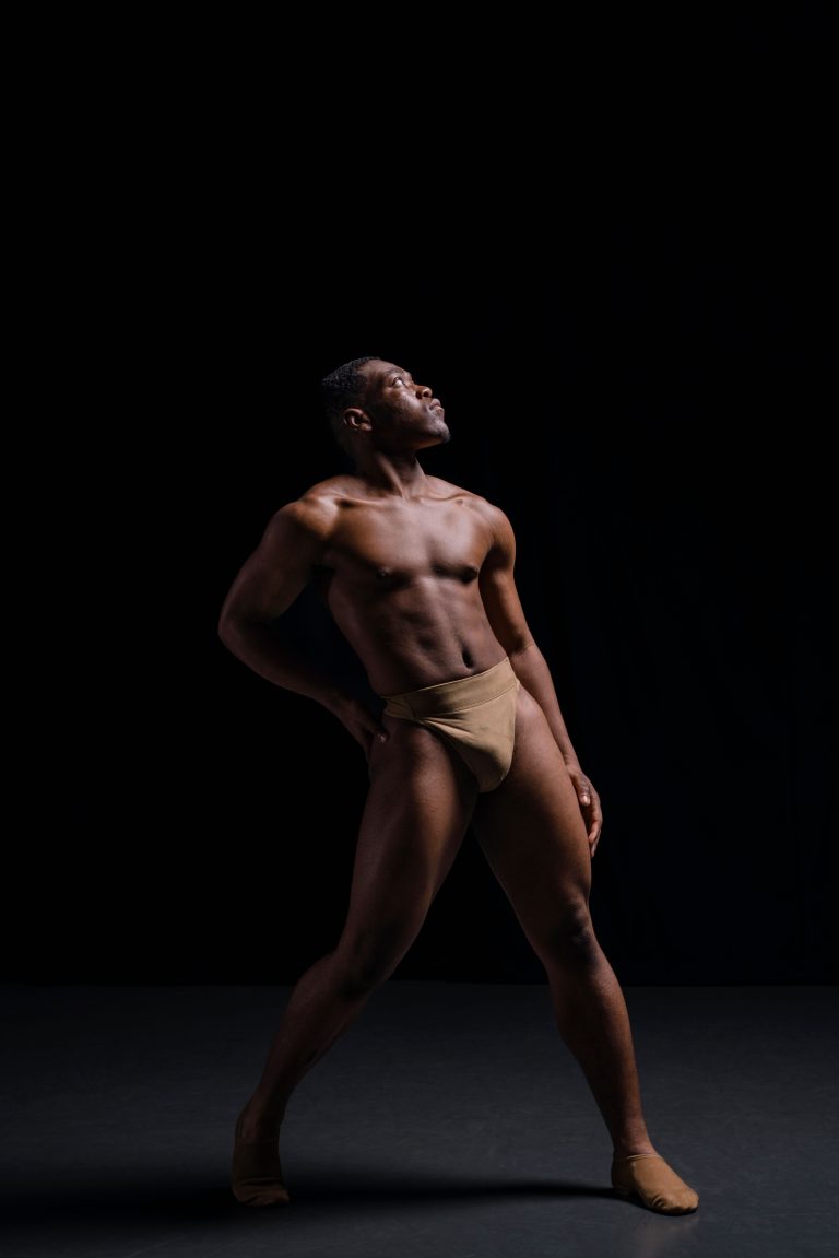 A male presenting dancer posing in his matching skin tone shades dancewear dance support belt ballet