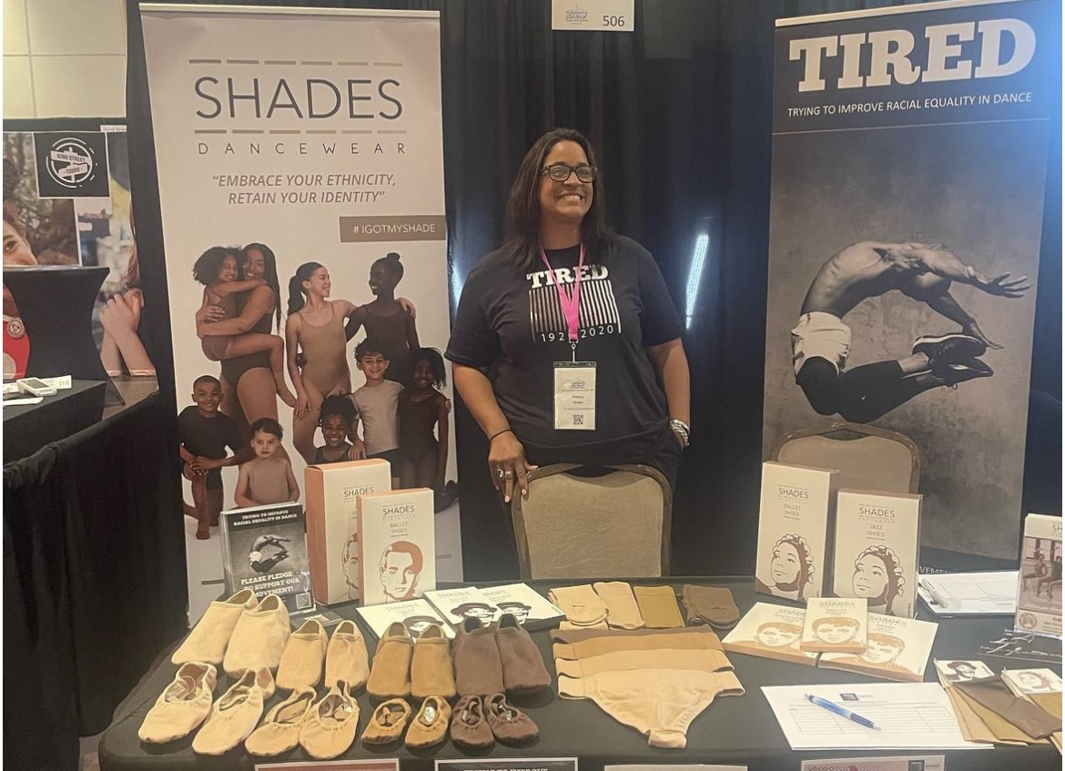 Photo shows Shades and TIRED co-founder Stacey Green, a mixed-race woman wearing a black TIRED t shirt, standing behind the Shades and TIRED stand at the Toronto Dance Teachers Expo 2024. There is a large Shades Dancewear banner to her left, and large TIRED banner to her right. Shades Dancewear products and packaging are laid across the table infront of her, along with a series of flyers.