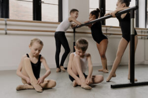 Image shows two children, one boy and one girl, sitting on the dance studio floor putting their matching skin tone ballet shoes on. There are 3 more children behind them leaning on the Ballet barre, 1 boy and 2 girls, also wearing matching skin tone dancewear