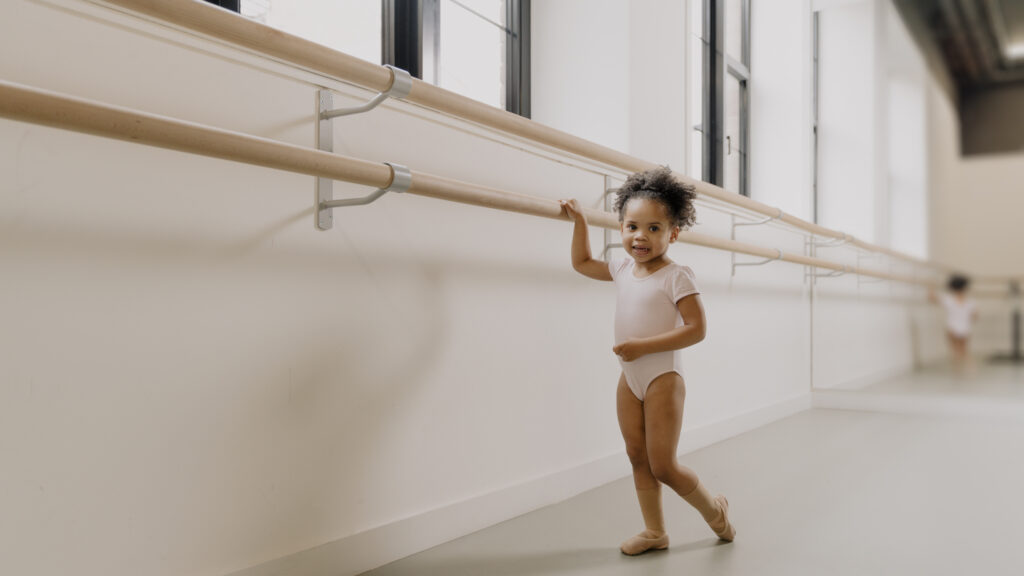 A young brown girl holding the Ballet barre in a dance studio, smiling. She is wearing a pink leotard and socks and ballet shoes that match the colour of her skin