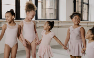 This photo shows 5 young dancers holding hands in a dance class studio, wearing various pink leotards.