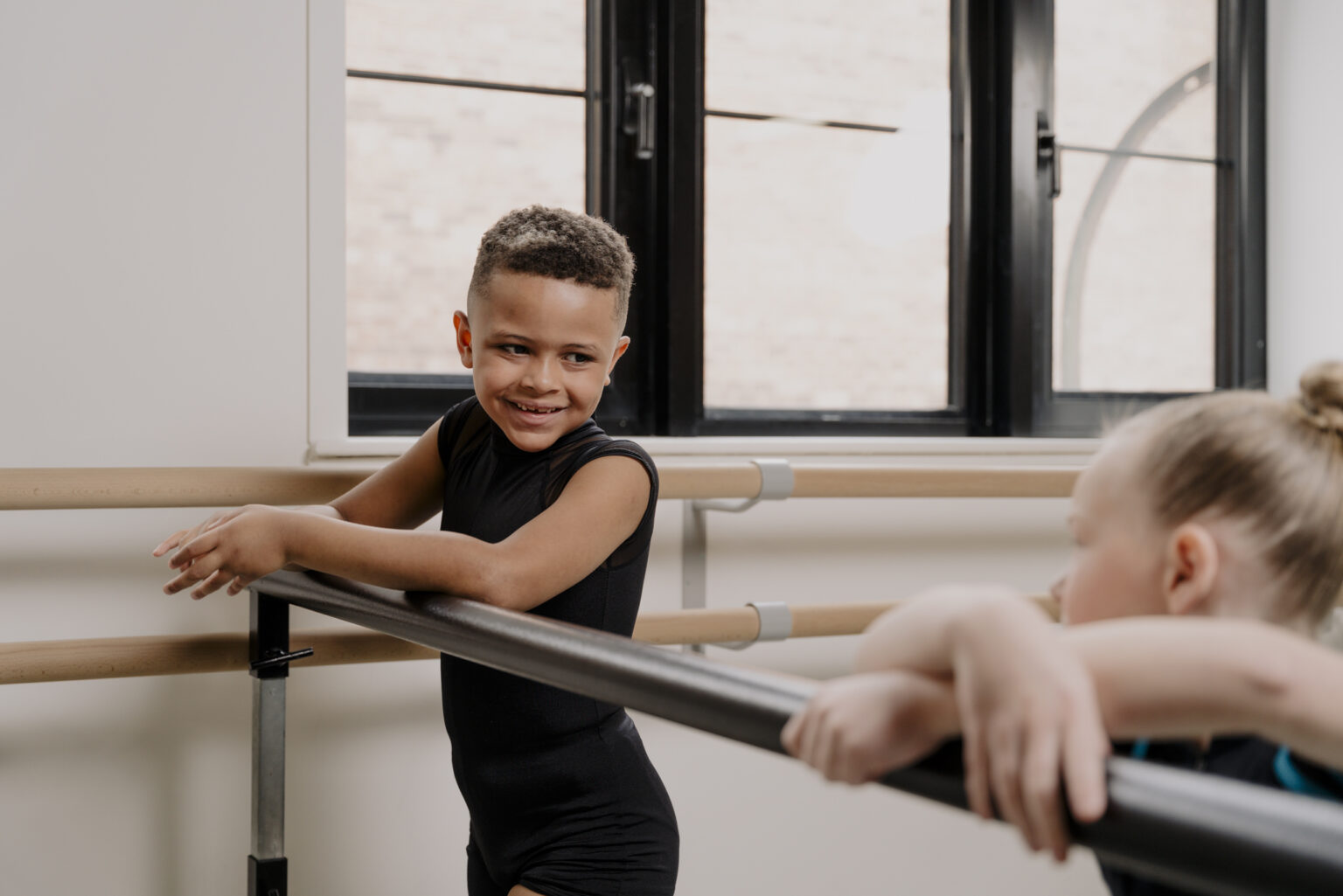 Picture shows one boy and one girl dancer at the Ballet barre smiling and looking at each other in a dance studio