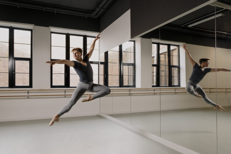 Picture shows a teenage boy in a dance studio jumping high in the air with one leg extended and the other bent at the knee