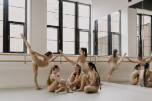 Picture shows a group of girls stretching in the dance studio by the Ballet barre, chatting and smiling at each other