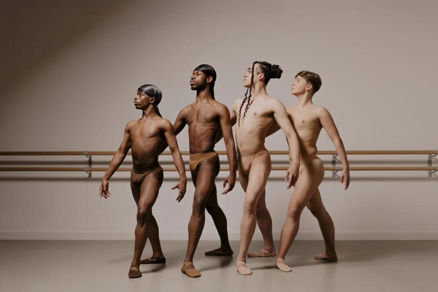 Four male-presenting ballet dancers of diverse skin tones and body types stand in a dance studio, posed in an elegant formation. Each dancer is wearing skin-tone dance belts and soft ballet shoes. They exude strength and grace, gazing in different directions with outstretched arms and poised postures. The background features ballet barres and a neutral-coloured wall.