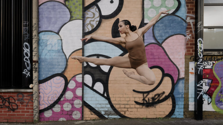 Picture shows a mixed race dancer jumping in the air with one leg straight out to the side and the other bent at the knee, foot pointed and almost touching the straight leg. Female dancer is wearing a brown leotard, matching skin tone tights and ballet shoes, hair in a ponytail. The dancer is jumping infront of multicoloured graffiti