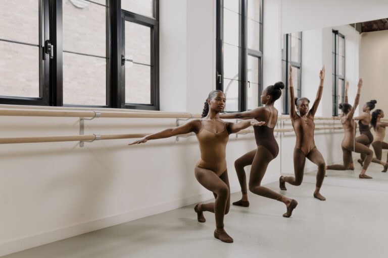 Three dancers in a studio performing exercises at a ballet barre. They are wearing flesh-tone leotards, tights, and jazz shoes that match their skin tones. The studio features large windows, natural light, and mirrors reflecting their movements.