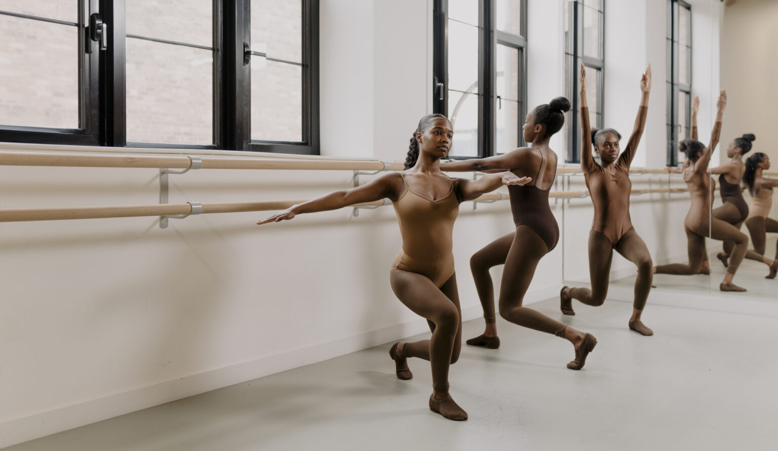 Three dancers in a studio performing exercises at a ballet barre. They are wearing flesh-tone leotards, tights, and jazz shoes that match their skin tones. The studio features large windows, natural light, and mirrors reflecting their movements.