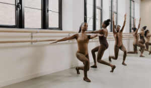 Three dancers in a studio performing exercises at a ballet barre. They are wearing flesh-tone leotards, tights, and jazz shoes that match their skin tones. The studio features large windows, natural light, and mirrors reflecting their movements.