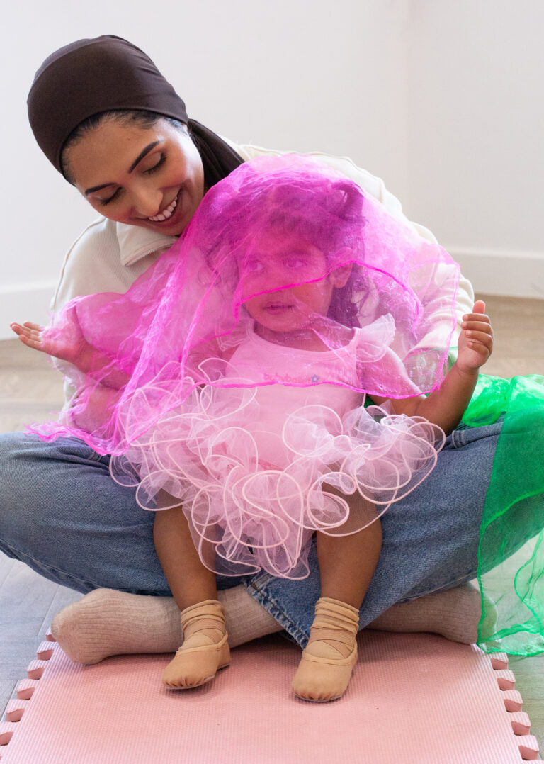 A smiling adult and young child share a joyful moment together on a soft mat. The adult, wearing a brown headscarf and a cozy top, lovingly embraces the child, who is dressed in a frilly pink ballet outfit and small ballet slippers. They playfully hold up colorful scarves, including a bright pink one that drapes over the child’s head, creating a whimsical and heartwarming scene.