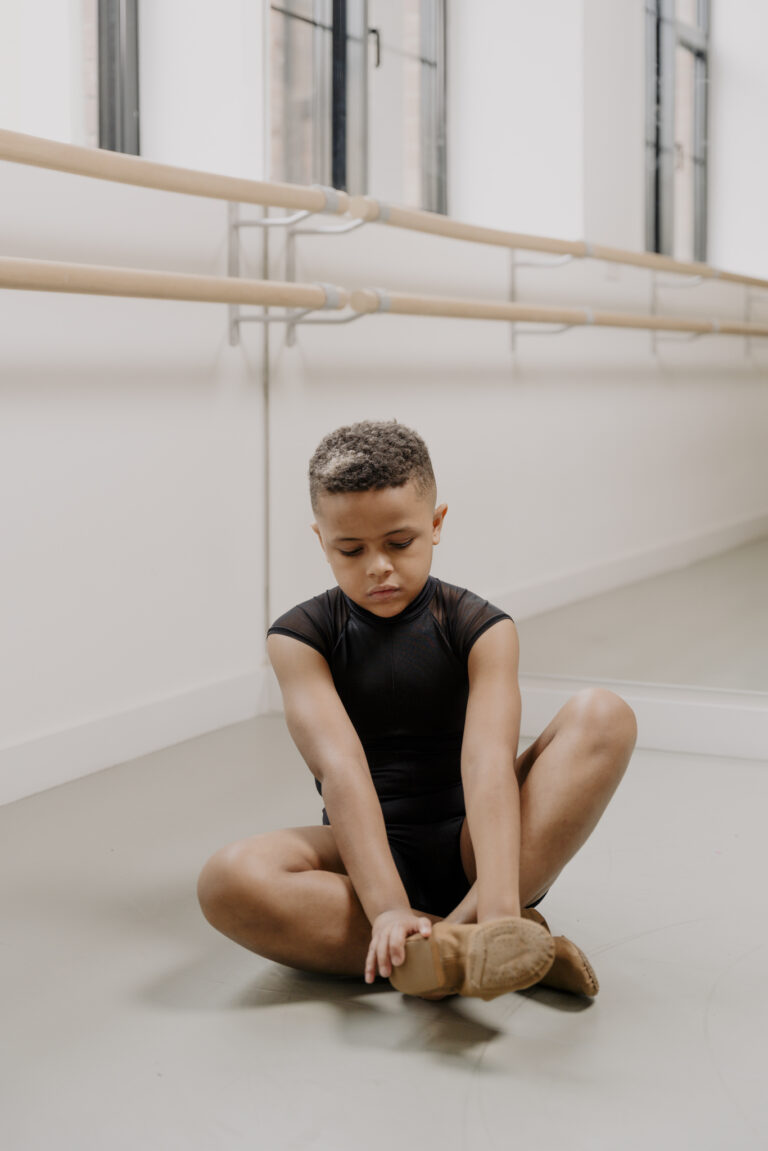 A young boy in a black dance leotard sits on the studio floor, stretching and adjusting his Jazz shoe, with ballet barres and mirrors in the background